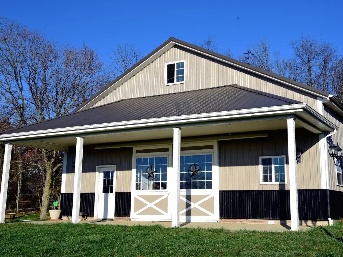 Tan metal pole building with a covered front porch, white posts, and star-accent doors