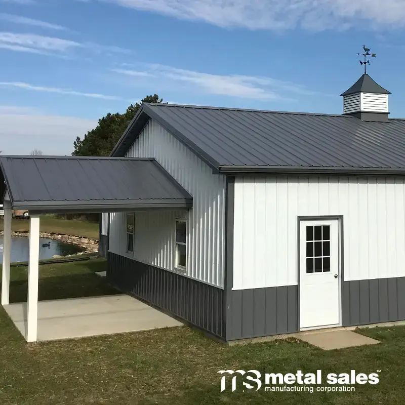 White and gray metal-sided building with cupola, covered patio and pond beyond