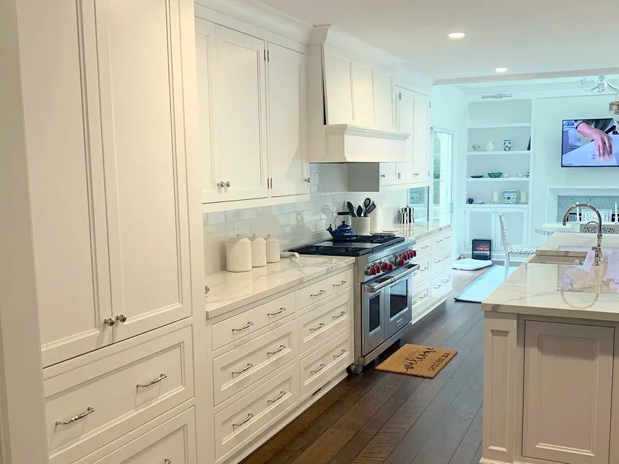 Long white kitchen with inset cabinets, range under a white hood and marble island