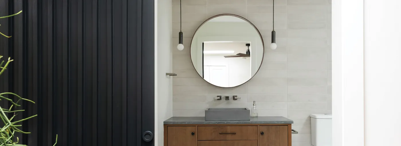 Bathroom with wood vanity, round mirror and pendant lights seen past a black door